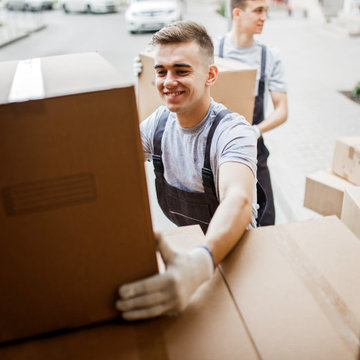 A young handsome smiling mover wearing uniform is reaching for the box while unloading the van full of boxes. House move, mover service.
